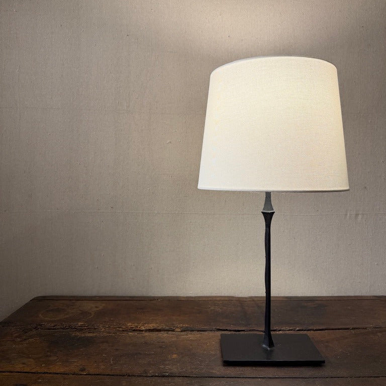 Cast iron table lamp with a pale tapered shade and square base on a wooden table.