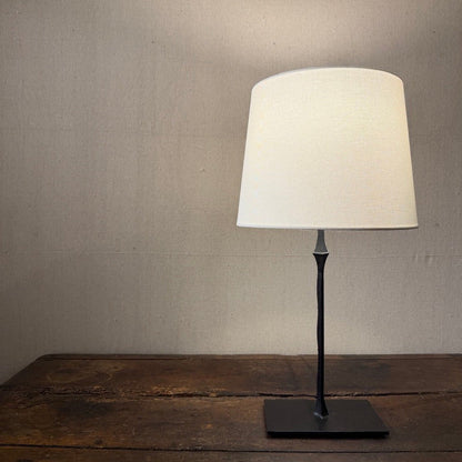 Cast iron table lamp with a pale tapered shade and square base on a wooden table.