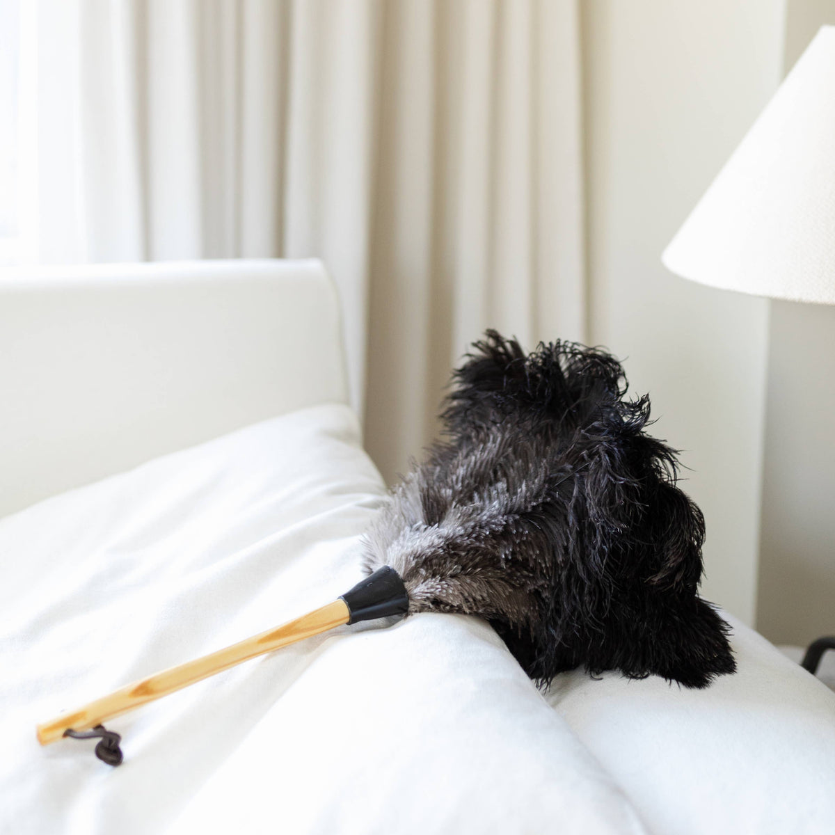 Black feather duster with a wooden handle resting on white bedding.