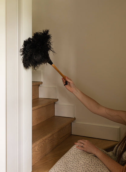 A 20-inch feather duster with dark feathers and a wooden handle, shown in use beside a staircase.